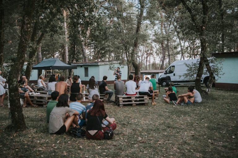 Ljudi sjede na paletama od drva i na podu u prirodi i slušaju edukaciju predavača na "Pandemonium Festivalu"